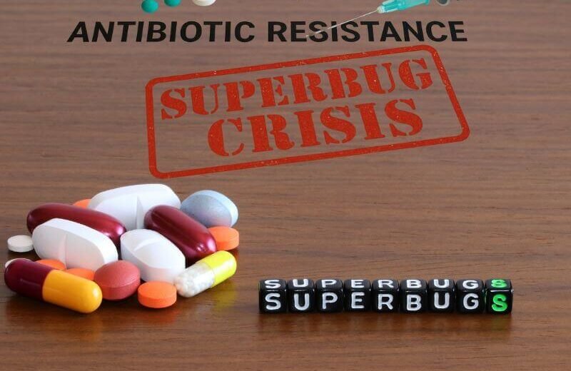 Realistic image showing medical supplies on a wooden surface with bold text "ANTIBIOTIC RESISTANCE", illustrating the superbug crisis.